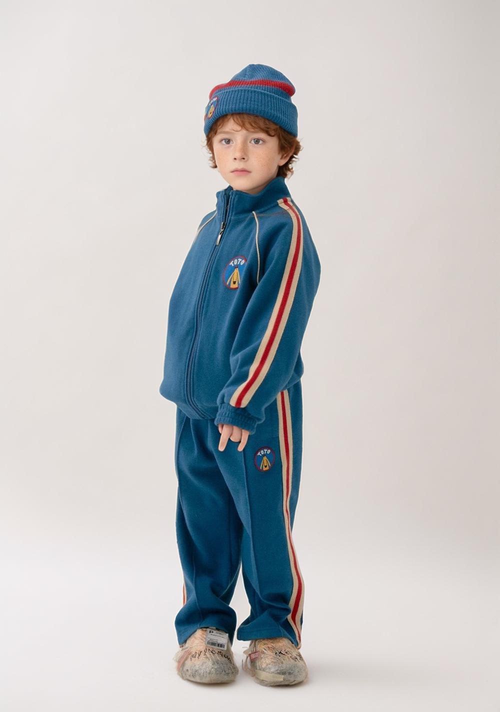 A Young Child Dressed In A Matching Blue Tracksuit And Beanie Stands Against A Plain Background