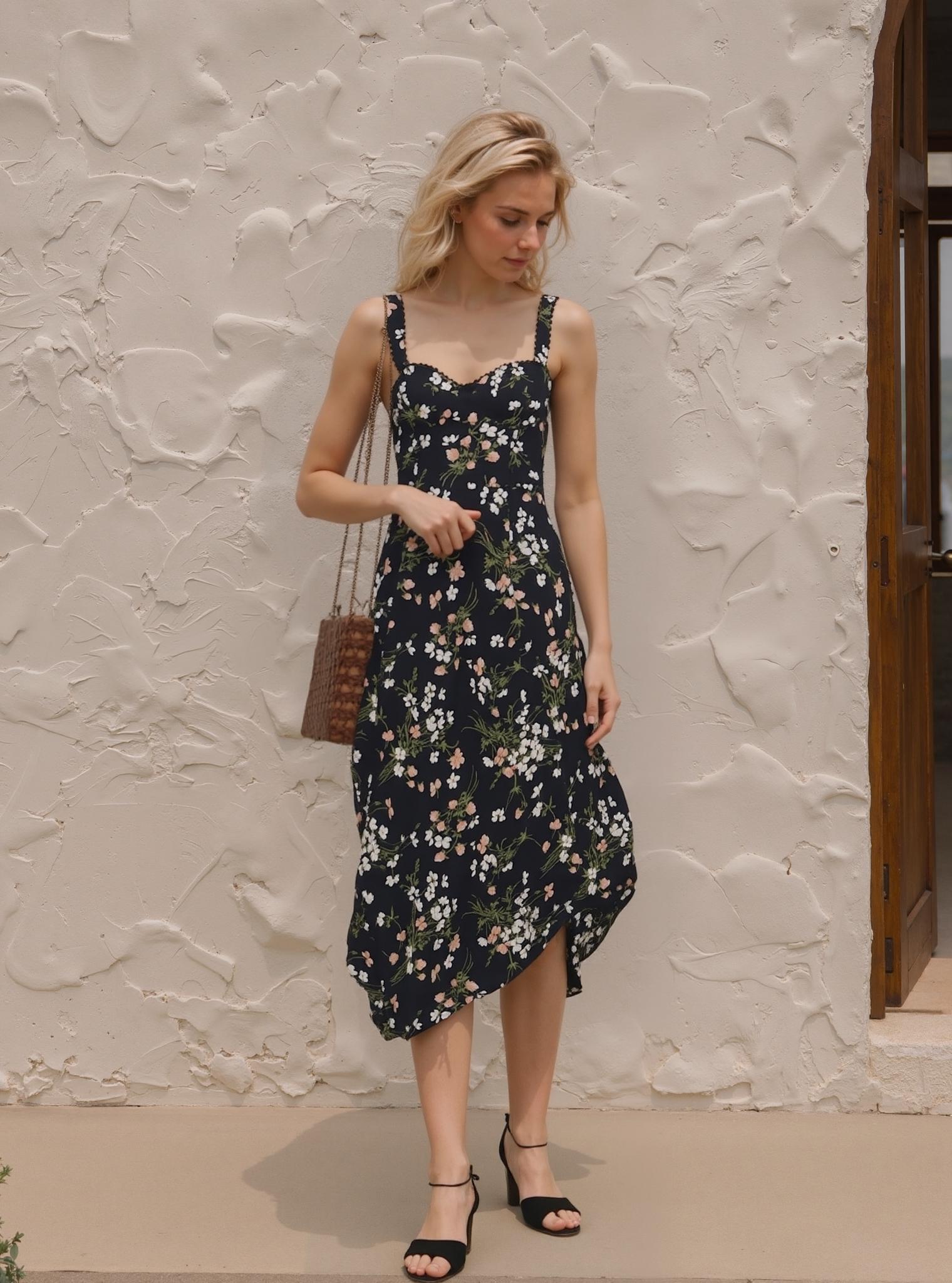 A Woman Stands Outdoors Against A Textured White Wall Wearing A Floral Dress And Black Heels While Carrying A Woven Purse