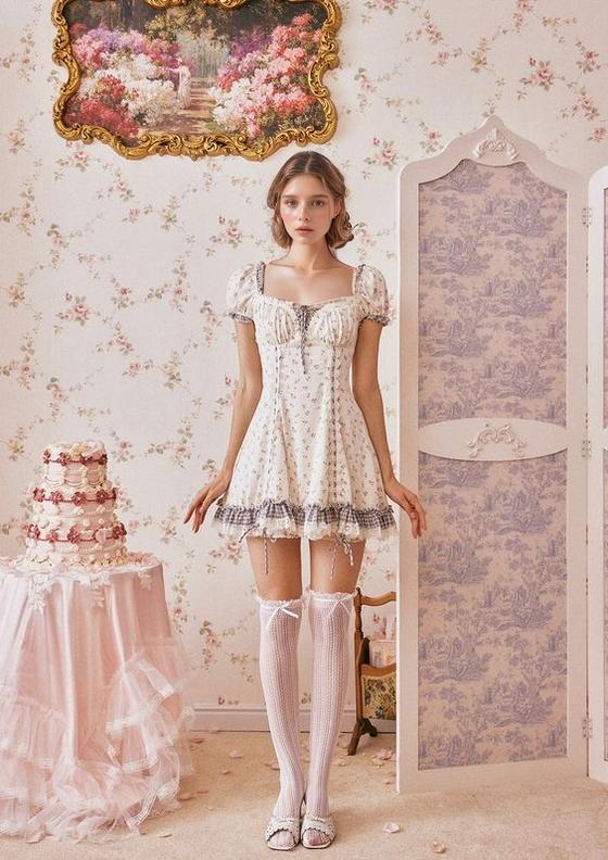 A Woman In A Floral Dress And Stockings Stands In A Vintage-Inspired Room Decorated With Floral Wallpaper And A Tiered Cake On A Table