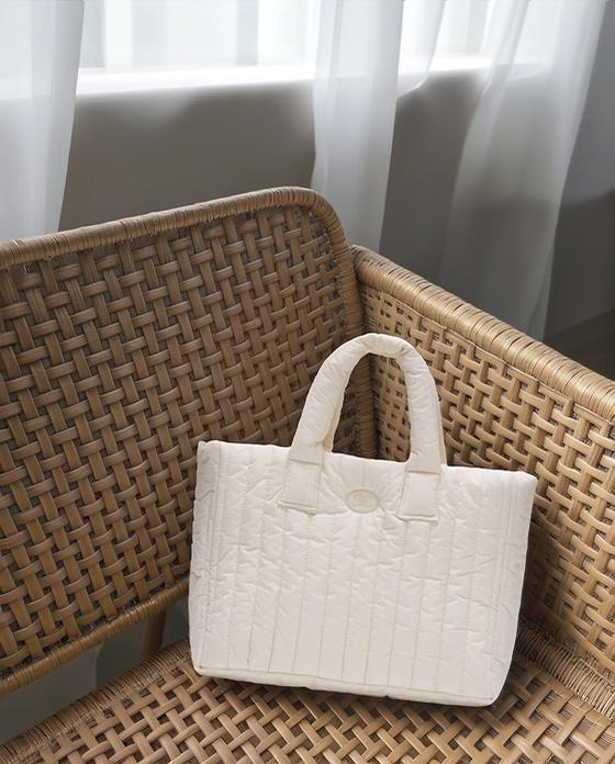 A White Quilted Bag Sits Elegantly On A Woven Chair By A Sunlit Window With Sheer Curtains