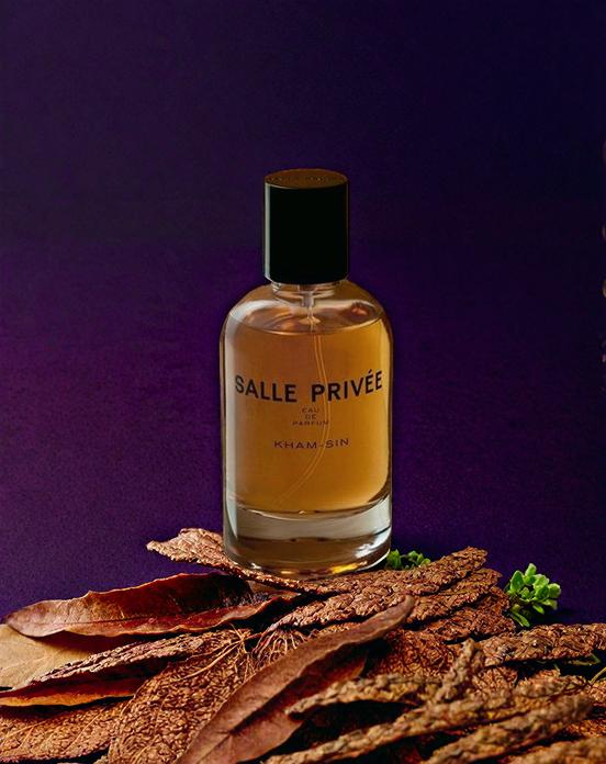 A Bottle Of Salle Privée Perfume Is Elegantly Displayed Against A Purple Backdrop With Dried Leaves At Its Base