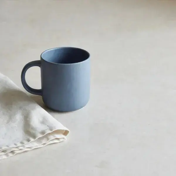 Matte blue ceramic mug on pale stone with a folded linen napkin and empty space on the right