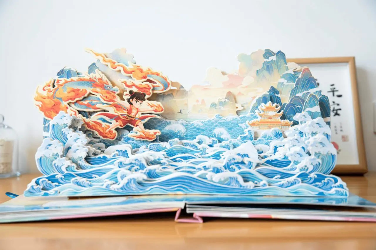 A pop-up paper book depicting Nezha and layered waves, presented front-on on a desk.