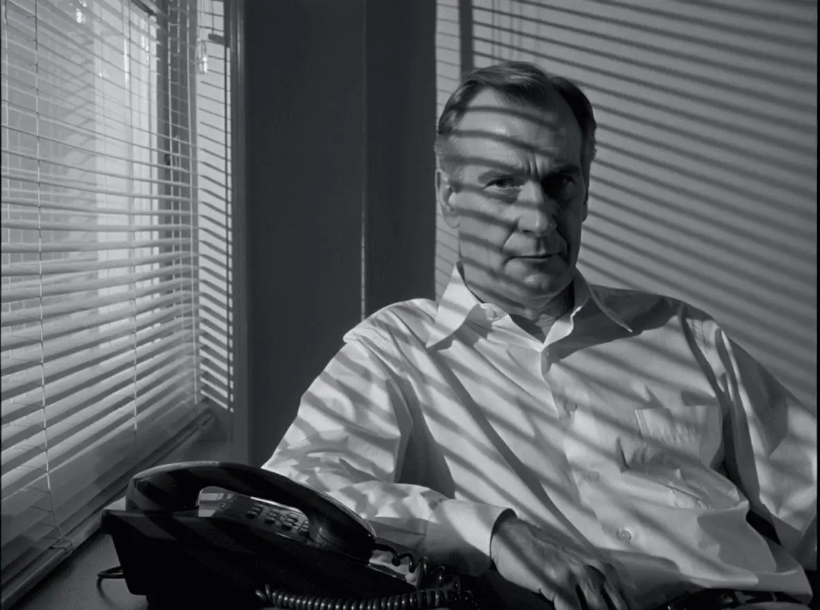 A seated detective and a desk telephone appear under striped light from window blinds.